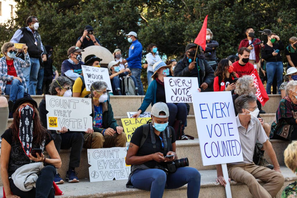 People sitting on benches at Frank H. Ogawa Plaza in Oakland, Calif., with signs supporting election integrity.